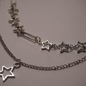 May include: A layered silver necklace featuring a chain with a dangling star pendant and a second chain with star-shaped charms. The necklace also has a chain with rectangular links and star-shaped charms. The jewelry is displayed on a light background.