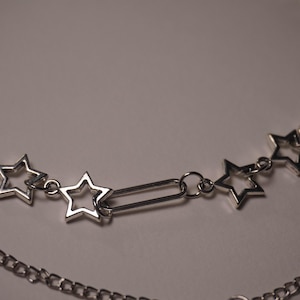 May include: A silver-toned, layered necklace featuring open star pendants and a delicate chain. The top layer has star-shaped charms connected by small links and a rectangular bar. The bottom layer is a simple chain.