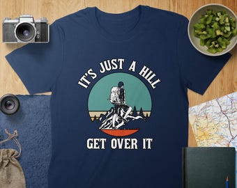 Op de afbeelding: Donkerblauw T-shirt met een retro-grafische afbeelding van een wandelaar op een berg. De tekst "It's just a hill get over it" staat op het shirt gedrukt.
