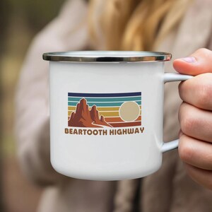 May include: White enamel mug featuring a retro design with a mountain range, sun, and horizontal stripes in shades of brown, blue, and orange. The text "Beartooth Highway" is printed below the graphic. The mug has a silver rim and a white handle.