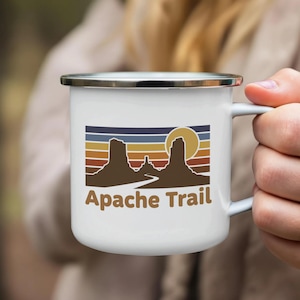 May include: White enamel camping mug with a black rim. The mug features a retro design with a landscape silhouette, a sun, and the text "Apache Trail" in brown. The design includes horizontal stripes in various colors.
