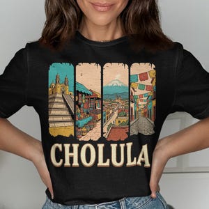 May include: Black t-shirt featuring a colorful graphic of Cholula, Mexico. The design includes illustrations of a pyramid, market, cityscape with a volcano, and a street with flags. The word "CHOLULA" is printed in large, tan letters.