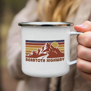 May include: White enamel mug with a silver rim featuring a retro design of mountains, a road, and a sun, with the text "BEARTOOTH HIGHWAY" in brown. The mug is held by a person.