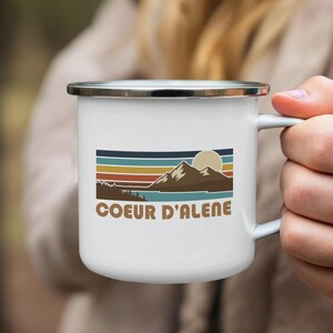 Puede incluir: Taza de esmalte blanco con borde plateado, con un diseño retro de montañas, un sol y rayas horizontales en tonos azules, naranjas y marrones. El texto "COEUR D'ALENE" está impreso debajo. La taza es sostenida por una persona.