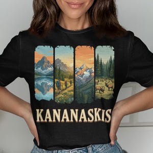 Pode incluir: Camiseta preta com um design de paisagem que retrata montanhas, árvores e um lago. O texto "KANANASKIS" é impresso em bege, com uma fonte de estilo vintage, na parte inferior da camiseta.