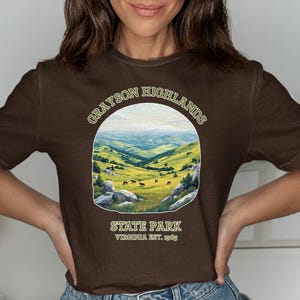 May include: A brown t-shirt featuring a scenic landscape design of Grayson Highlands State Park in Virginia. The design includes rolling green hills, blue mountains, and the text "Grayson Highlands State Park Virginia Est. 1965."