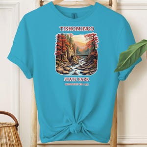 May include: A turquoise blue t-shirt with a graphic of a scenic view of a bridge over a river in a forest. The text "Tishomingo State Park Mississippi Est. 1935" is printed below the graphic.