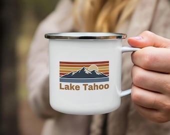 May include: White enamel mug with a black rim and handle. The mug features a retro design with a mountain range, sun, and the words "Lake Tahoe" in brown. The design is set against a backdrop of horizontal stripes in shades of brown and orange.