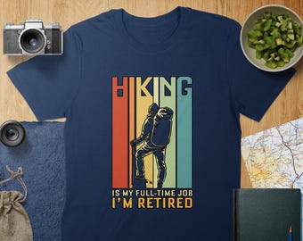 Op de afbeelding: Een marineblauw t-shirt met een vintage-stijl afbeelding van een wandelaar met een rugzak. De tekst "Hiking is my full-time job I'm retired" staat onder de afbeelding.