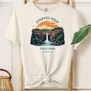 Può includere: Una maglietta bianca con un design grafico di una cascata in un parco statale. La grafica dice "Starved Rock State Park, Illinois Est. 1911".
