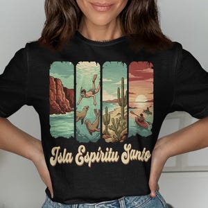 May include: Black t-shirt with a colorful graphic of Isla Espíritu Santo scenes. The design includes a cliffside, a diver with seals, a beach with cacti, and kayakers at sunset. The text "Isla Espíritu Santo" is in a vintage font.
