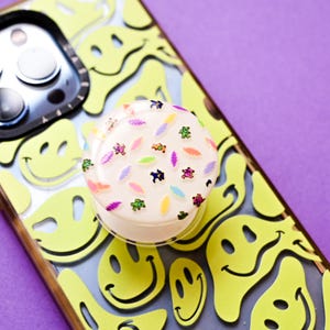 May include: A phone case with a yellow smiley face pattern and a white pop socket. The pop socket is decorated with colorful confetti. The phone case is on a purple background.