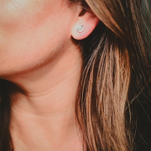 May include: A close-up of a silver stud earring with a small, delicate leaf design. The earring is in a person's ear, with the focus on the jewelry. The background is blurred, highlighting the earring's details.