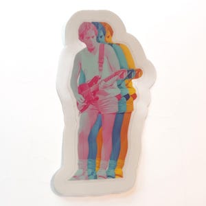May include: A sticker featuring a multi-colored image of a person playing a red electric guitar. The image has a retro, layered effect, with the figure appearing in shades of pink, blue, yellow, and orange. The sticker has a white border.