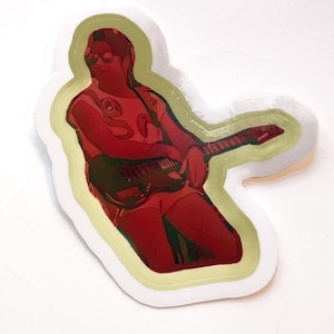 May include: A sticker featuring a red and black illustration of a person playing a guitar. The person is wearing sunglasses and a white shirt with a snake design. The sticker has a white border with a light green outline.