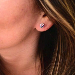 May include: A close-up of a silver stud earring with a colorful, circular design. The design features a red, blue, and white graphic. The earring is in a person's ear, with their skin and hair visible.