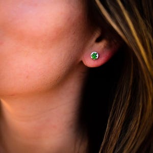 May include: A small, round stud earring featuring a green bear design on a white background. The earring is set against the backdrop of a person's ear and skin, with a blurred background.
