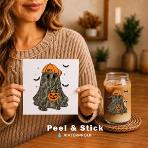 May include: A square sticker featuring a ghost wearing an orange baseball cap and holding an orange jack-o'-lantern bucket, with a camouflage pattern on the ghost's body, surrounded by small black bats and stars on a white background.