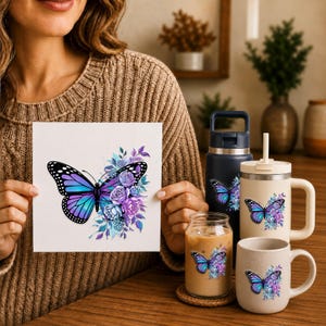 May include: A square print, a dark blue water bottle, a cream-colored tumbler with a handle and straw, and a cream-colored mug, all featuring a graphic of a blue and purple butterfly surrounded by purple and blue flowers and foliage.