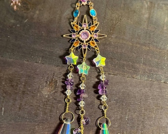 May include: A gold-toned sunburst charm with iridescent purple, blue, and orange crystals hangs from a chain with iridescent purple and clear crystal beads and two iridescent crystal teardrop pendants.