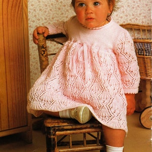 Könnte beinhalten: Ein kleines Kind in einem rosa Strickkleid mit Spitzenmuster sitzt auf einem Holzstuhl. Das Kind trägt weiße Socken und weiße Schuhe.