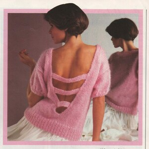 May include: A pink knitted sweater with a large open back design featuring criss-cross straps. The sweater has short sleeves and a relaxed fit.