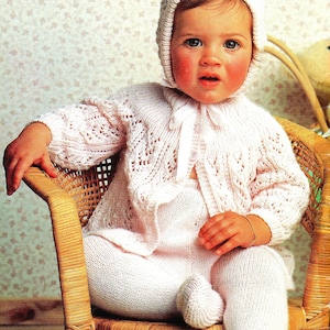May include: A baby wearing a white knitted hat, cardigan, and pants. The baby is sitting in a wicker chair.