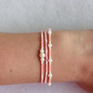 Może przedstawiać: Trzyrzędowa bransoletka z różowymi i białymi koralikami z perłowymi akcentami. Bransoletka jest owinięta wokół nadgarstka, prezentując delikatny design i paletę kolorów. Koraliki są małe i równomiernie rozmieszczone.