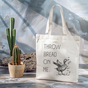 Puede incluir: Bolsa de tela blanca de lona con texto negro que dice "THROW BREAD ON ME." Una ilustración en blanco y negro de un pato con migas de pan alrededor está en la bolsa.