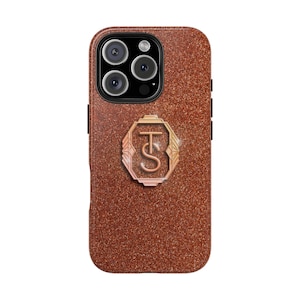 Puede incluir: Una funda de iPhone marrón brillante con un emblema metálico con las letras "TS". La funda tiene un módulo de cámara con tres lentes y un flash. La funda está diseñada para proteger el teléfono y añadir un toque de estilo.