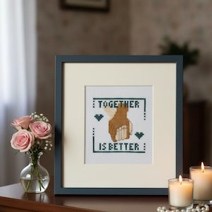 May include: Framed cross-stitch art with the text "TOGETHER IS BETTER" and two hands clasped. The artwork is in a dark blue frame, displayed on a wooden surface with candles and flowers. The embroidery is sold unframed.