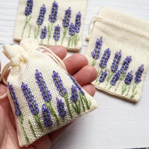 May include: Three small, off-white drawstring pouches made of a textured fabric, each featuring an embroidered design of purple lavender sprigs with green stems. The pouches have a simple closure with matching off-white drawstrings at the top.