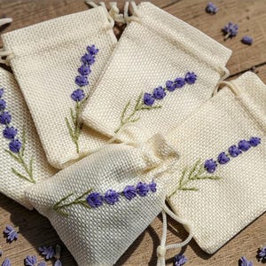 Puede incluir: Cinco pequeñas bolsas beige con cordón, bordadas con ramitas de lavanda morada. Las bolsas están dispuestas sobre una superficie de madera, rodeadas de capullos de lavanda sueltos y unos cuantos tallos de flores amarillas. Las bolsas son de tela texturizada.