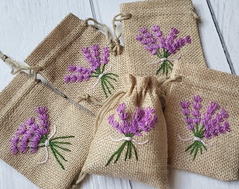 May include: A collection of burlap drawstring bags, each adorned with an embroidered lavender sprig. The purple flowers are detailed with green stems and tied with a white ribbon. The bags are a natural beige color.