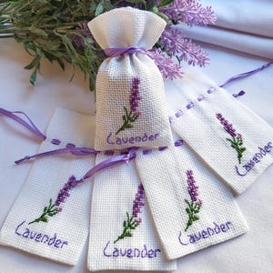 Könnte beinhalten: Weiße Leinenbeutel mit lila Lavendelzweigen und dem Wort "Lavender" bestickt. Ein Beutel ist ein Kordelzugbeutel, die anderen sind flach. Lila Bandakzente. Ein Lavendelzweig ist im Hintergrund.