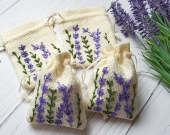 May include: Four small, cream-colored drawstring bags with embroidered lavender sprigs in purple and green. The bags are arranged with a sprig of lavender and green leaves in the background. The bags are made of a woven fabric.