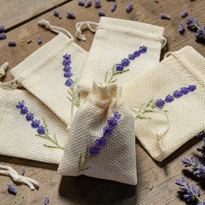 Puede incluir: Cinco pequeños saquitos de lino beige con bordados de ramitas de lavanda morada están dispuestos sobre una superficie de madera. Brotes y ramitas de lavanda dispersos rodean los saquitos, creando una presentación natural y aromática.