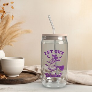May include: A clear glass can-shaped tumbler with a pale pink lid and a silver straw. The tumbler has a purple graphic of a witch on a broom with the text "LET GET WICKED". A fun Halloween-themed drinking glass.