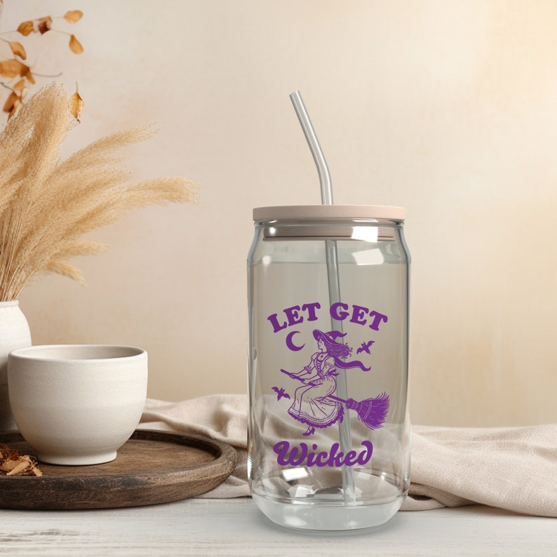 May include: A clear glass can-shaped tumbler with a pale pink lid and a silver straw. The tumbler has a purple graphic of a witch on a broom with the text "LET GET WICKED". A fun Halloween-themed drinking glass.