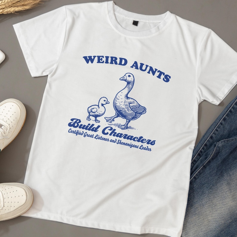 May include: White t-shirt with a blue graphic of two geese and the text "Weird Aunts Build Characters Certified Great Listener and Shenanigans Leader."