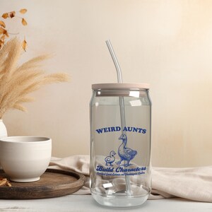 May include: A clear glass can with a pink lid and a blue and white design that says "Weird Aunts Build Characters Certified Great Listeners and Shameless Leaders". The can has a clear straw sticking out of the top.