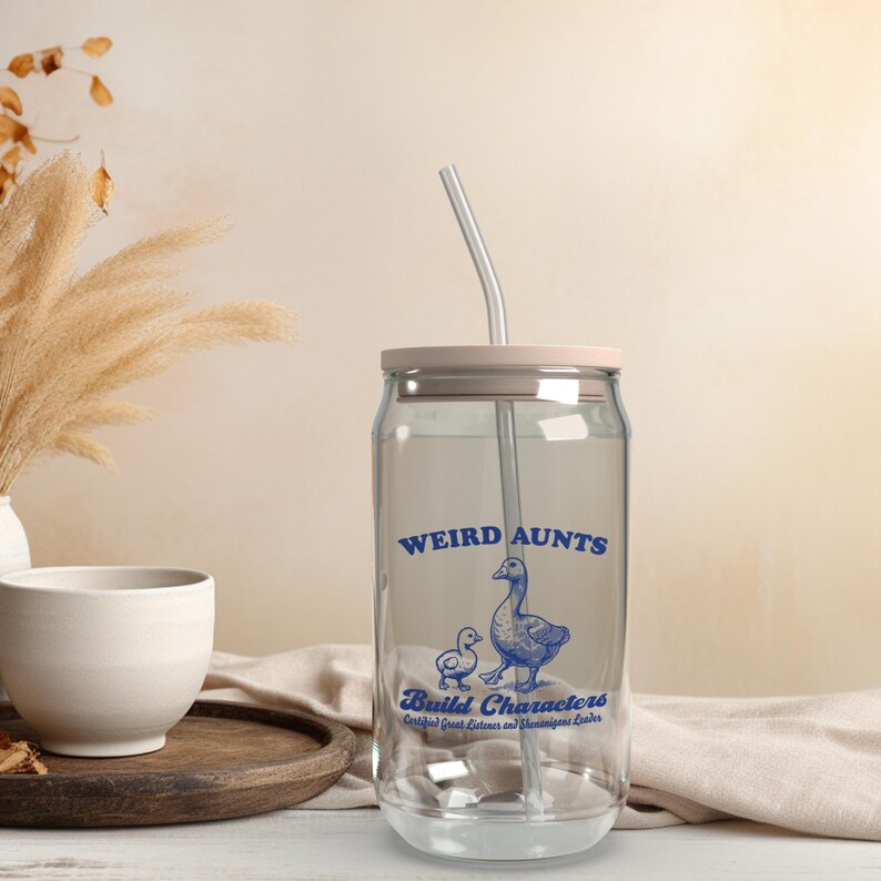 May include: A clear glass can with a pink lid and a blue and white design that says "Weird Aunts Build Characters Certified Great Listeners and Shameless Leaders". The can has a clear straw sticking out of the top.
