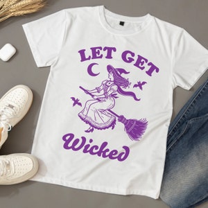 May include: White t-shirt featuring a purple witch graphic on a broomstick, with the text "LET GET Wicked". The witch is depicted in a hat and dress. The shirt is styled with white trainers and blue jeans.