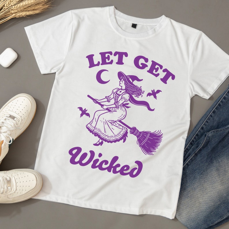 May include: White t-shirt featuring a purple witch graphic on a broomstick, with the text "LET GET Wicked". The witch is depicted in a hat and dress. The shirt is styled with white trainers and blue jeans.