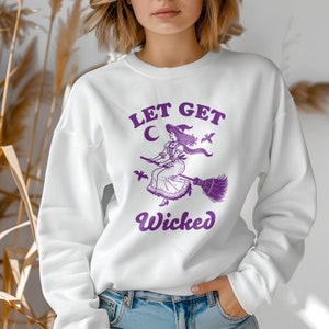 May include: White sweatshirt featuring a purple graphic of a witch on a broom, with the words "LET GET Wicked" above and below. The witch illustration includes a hat and dress, with bats flying nearby. A crescent moon is also visible.
