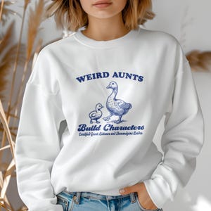 May include: White sweatshirt with a blue graphic of two geese and the text "Weird Aunts Build Characters Certified Great Listener and Shenanigans Leader."