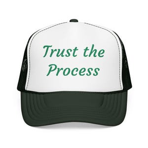 Peut inclure: Casquette de camionneur avec un panneau avant blanc et une visière et un dos en filet vert foncé. Le texte "Trust the Process" est imprimé en vert sur le devant de la casquette.