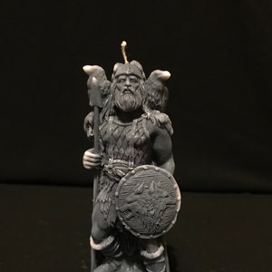 Könnte beinhalten: Eine graue Kerze in Form einer Wikingerfigur, die einen Speer und einen Schild hält. Der Schild hat ein Wolfsdesign. Die Figur hat einen langen Bart, einen Helm und zwei Rabenfiguren auf den Schultern. Auf der Basis der Kerze steht "ODIN".