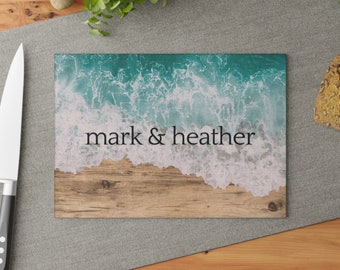 Könnte beinhalten: Rechteckiges Schneidebrett mit Strandmotiv. Die obere Hälfte zeigt einen türkisfarbenen Ozean mit weißen Wellen, der in einen Sandstrand übergeht. Die Namen "mark & heather" sind in Schwarz gedruckt.