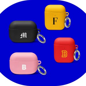 Könnte beinhalten: Vier Airpod-Hüllen mit verschiedenen farbigen Silikonbezügen und Buchstabeninitialen. Die Hüllen sind schwarz, gelb, rot und rosa. Jede Hülle hat einen silbernen Metallring, der oben angebracht ist.